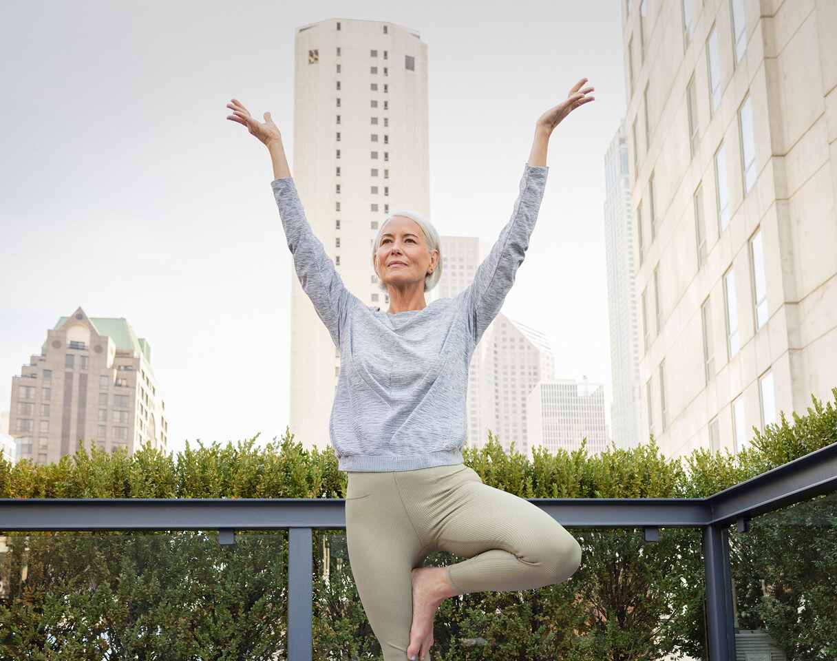 Woman standing on one foot with arms lifted skyward on an outdoor terrace with city buildings in the background.