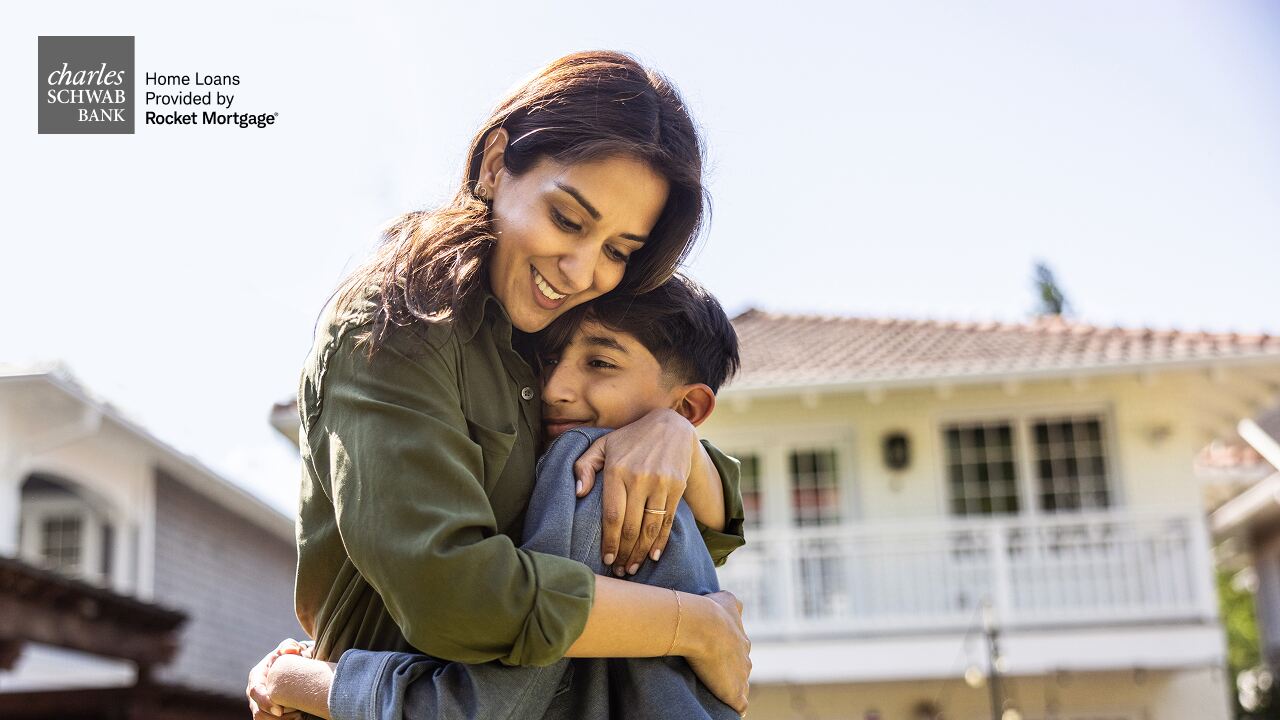 A woman hugs a smiling child outdoors in front of a home on a sunny day