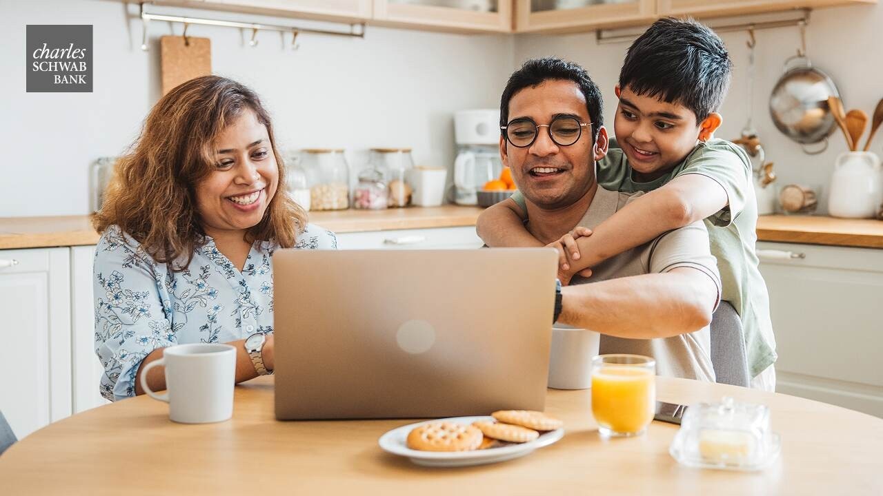 A family is sitting at the kitchen table, looking at a laptop screen.