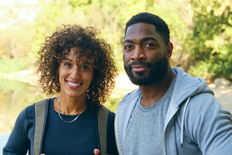A couple standing outdoors near a lake, smiling at the camera