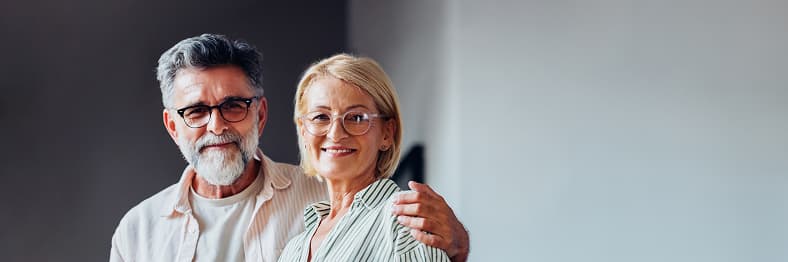 A smiling couple with glasses standing indoors, with the man’s arm around the woman's shoulder.