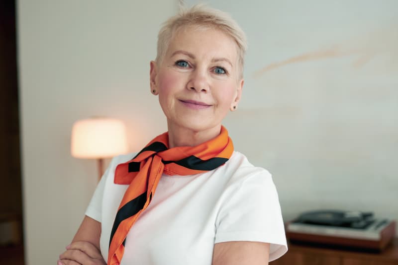A woman with short blonde hair wearing an orange scarf smiles softly.