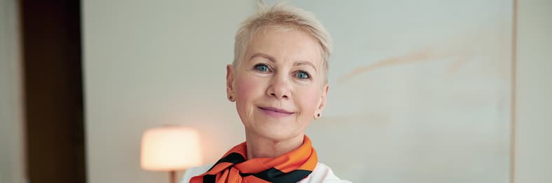 A woman with short blonde hair wearing an orange scarf smiles softly.