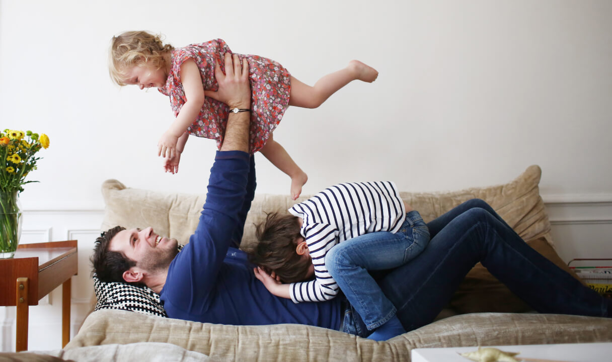 A father lies on a couch holding his small daughter above him, while another child lies on him.